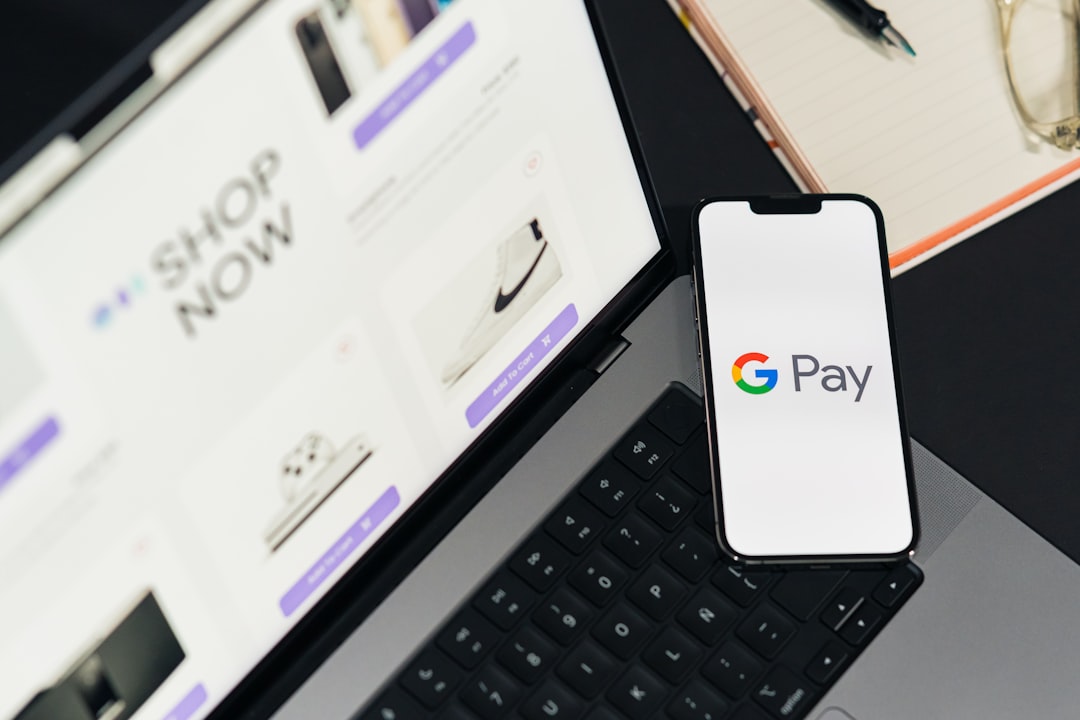 Google Pay displayed on a phone screen. Top-down view of a workspace. Desk with a laptop showing ecommerce on the screen, along with a mobile phone and notepad.